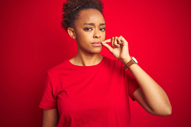 Fiatal gyönyörű afro-amerikai nő afro hajú át elszigetelt piros háttér száj és az ajkak zárva a zip ujjal. Titkos és csendes, tabu beszél - Fotó, kép