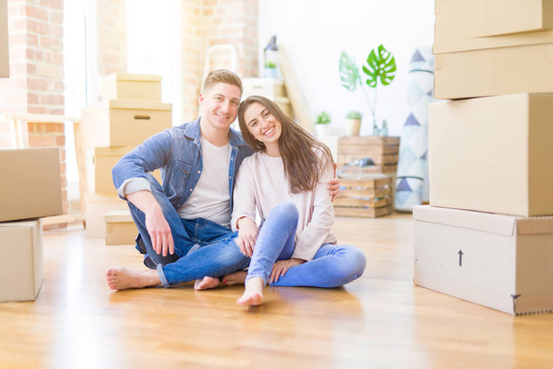 Joven hermosa pareja relajándose sentada en el suelo alrededor de cajas de cartón en casa, sonriendo feliz mudanza a una nueva casa
 - Foto, Imagen