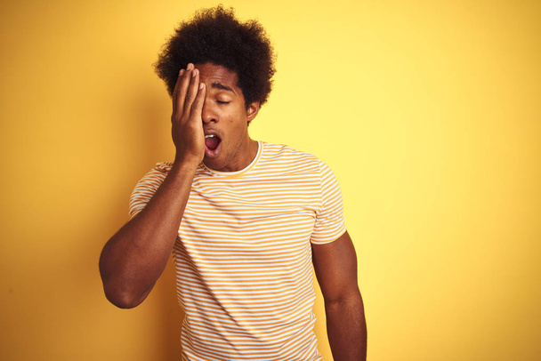 Amerikaanse man met Afro haar dragen gestreepte t-shirt staande over geïsoleerde gele achtergrond geeuwen moe bedekt half gezicht, oog en mond met de hand. Gezicht doet pijn. - Foto, afbeelding