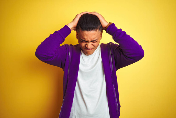 Mladý brazilský muž na purpurovém tričku, který stál nad izolovaným žlutým pozadím, trpí zoufalými bolestmi a tlakem, protože bolest a migréna. Ruce na hlavu. - Fotografie, Obrázek