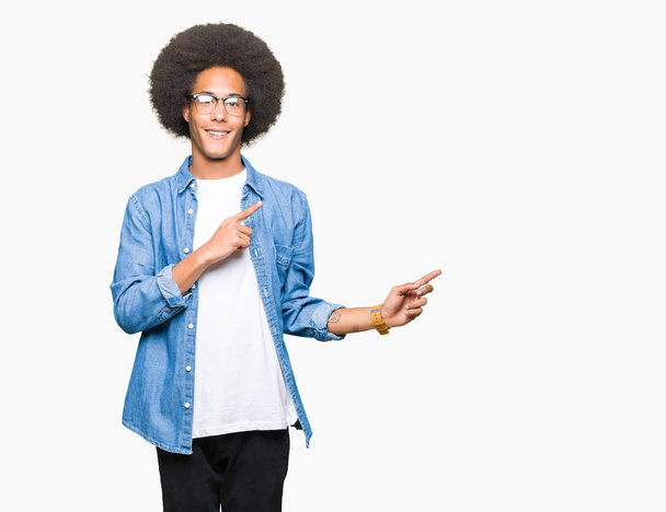 Young african american man with afro hair wearing glasses smiling and looking at the camera pointing with two hands and fingers to the side. - Foto, afbeelding