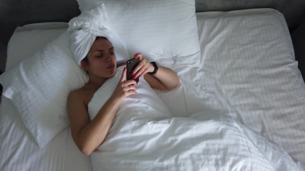 Vista dall'alto giovane femmina svegliarsi nel letto sotto coperta. Ragazza carina controlla il suo smartphone prima cosa al mattino
. - Filmati, video