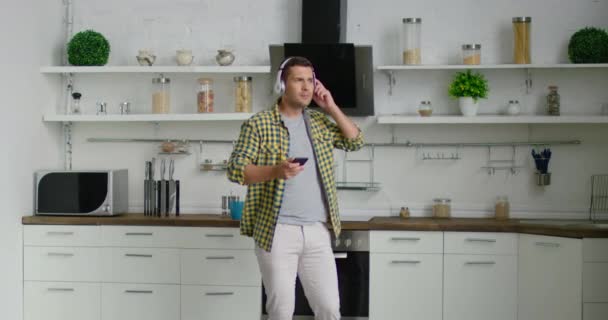 Handsome man is listening music in kitchen, using gadget and wireless headset - Filmmaterial, Video