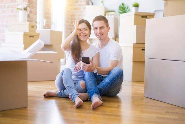 schönes Paar auf dem Boden sitzend mit Smartphone im neuen Zuhause um Pappkartons herum mit glücklichem Gesicht lächelnd ok Zeichen mit Hand auf Auge durch Finger schauend - Foto, Bild