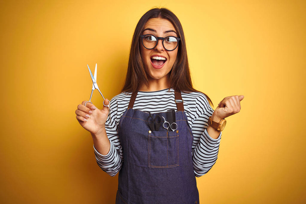 Young beautiful hairdresser woman holding scissors standing over isolated yellow background screaming proud and celebrating victory and success very excited, cheering emotion - Photo, Image