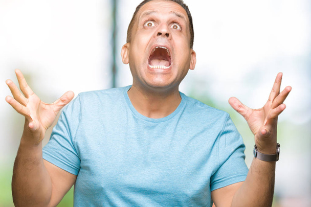 Homem árabe de meia idade vestindo camiseta azul sobre fundo isolado celebrando louco e espantado pelo sucesso com os braços levantados e olhos abertos gritando animado. Conceito de vencedor
 - Foto, Imagem