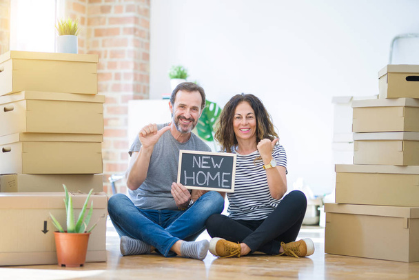 Középkorú vezető pár ül a padlón gazdaság táblára költözött egy új otthont mutató, és mutatja hüvelykujjával fel az oldalsó boldog arc mosolygós - Fotó, kép