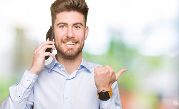 junger gutaussehender Mann spricht auf Smartphone und zeigt mit dem Daumen zur Seite mit glücklichem Gesicht lächelnd - Foto, Bild