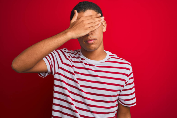 junger gutaussehender arabischer Mann mit gestreiftem T-Shirt über isoliertem rotem Hintergrund, der die Augen mit der Hand bedeckt und ernst und traurig aussieht. Versteck- und Ablehnungskonzept - Foto, Bild