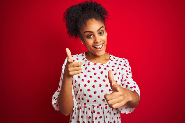Afro-amerikai nő visel divat, fehér ruhában állva az elszigetelt vörös háttér mutató ujjak a kamera boldog és vicces arc. Jó energia és vibrafon. - Fotó, kép