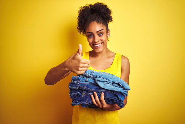Jonge African American vrouw Holding stapel jeans over geïsoleerde gele achtergrond gelukkig met grote glimlach doen OK teken, duim omhoog met de vingers, uitstekende teken - Foto, afbeelding