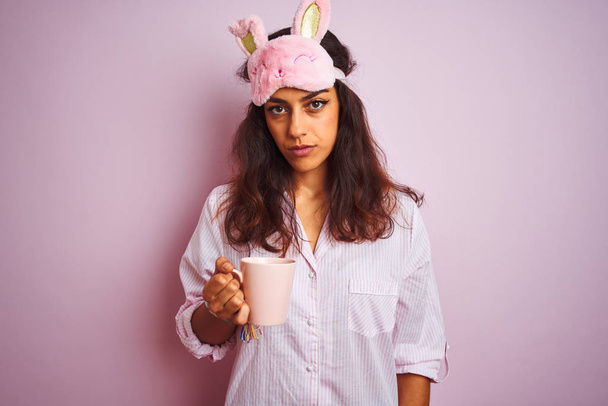 Young woman wearing pajama and sleep mask drinking coffee over isolated pink background with a confident expression on smart face thinking serious - Photo, Image
