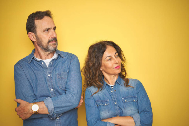 Beautiful middle age couple together standing over isolated yellow background looking to the side with arms crossed convinced and confident - Foto, immagini
