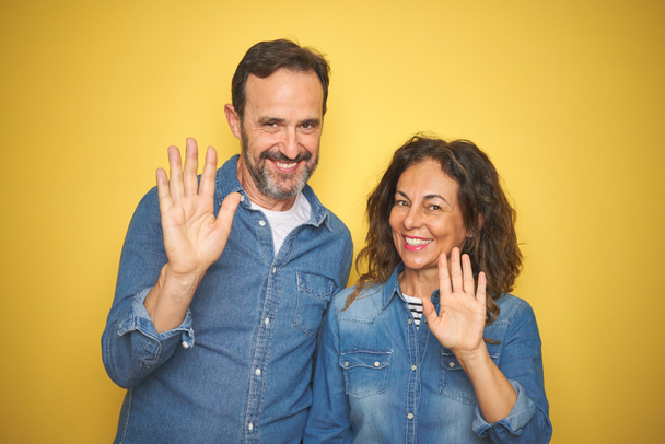 Bella coppia di mezza età insieme in piedi sopra isolato sfondo giallo rinuncia dicendo ciao felice e sorridente, amichevole gesto di benvenuto
 - Foto, immagini