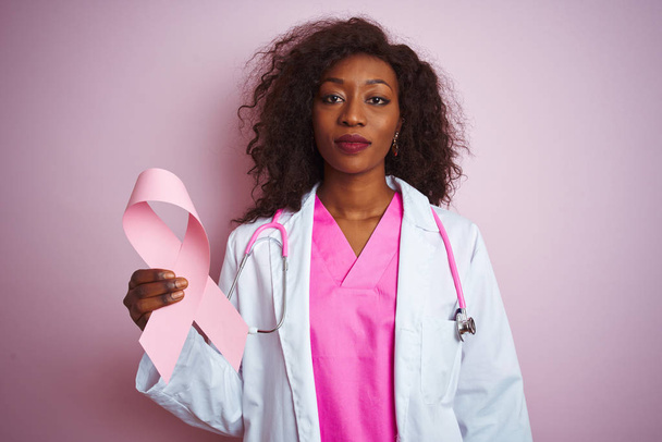 孤立したピンクの背景の上に癌のリボンを持つ若いアフリカ系アメリカ人の医師の女性は、真剣に考えるスマートな顔に自信のある表情で - 写真・画像