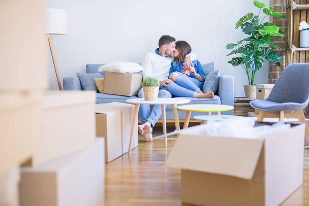 Young beautiful couple sitting on the sofa kissing at new home around cardboard boxes - Photo, Image