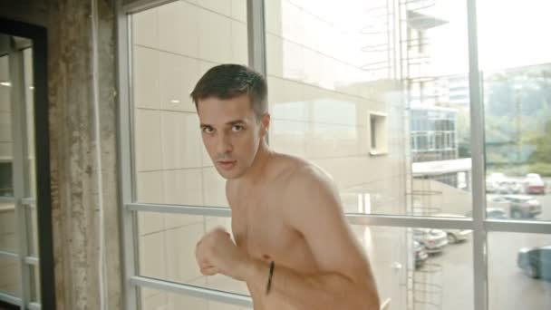 An athletic shirtless man boxer standing by the window and boxing in front of the camera - Séquence, vidéo