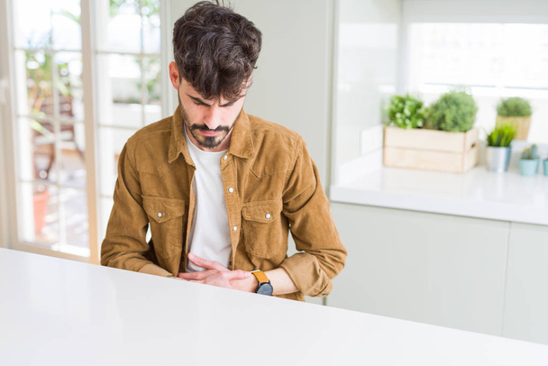 Young man wearing casual jacket sitting on white table with hand on stomach because nausea, painful disease feeling unwell. Ache concept. - Photo, Image