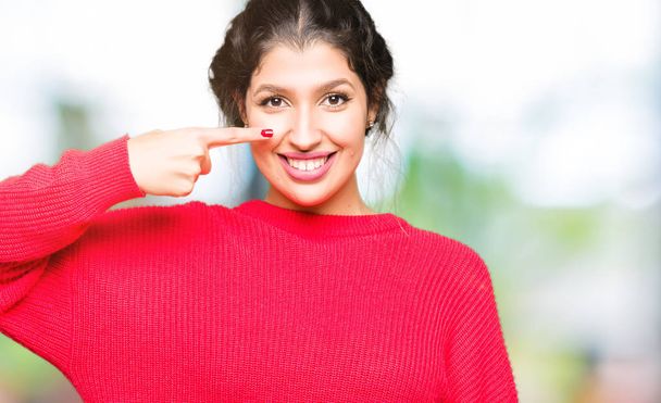 Kırmızı kazak ve topun giyen genç güzel kadın yüz ve burun el parmağı ile işaret, neşeli gülümseyen - Fotoğraf, Görsel