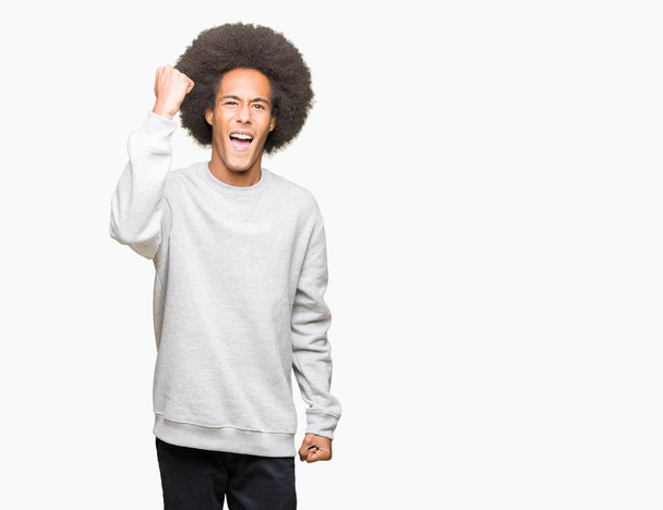 Young african american man with afro hair wearing sporty sweatshirt angry and mad raising fist frustrated and furious while shouting with anger. Rage and aggressive concept. - Foto, Imagem