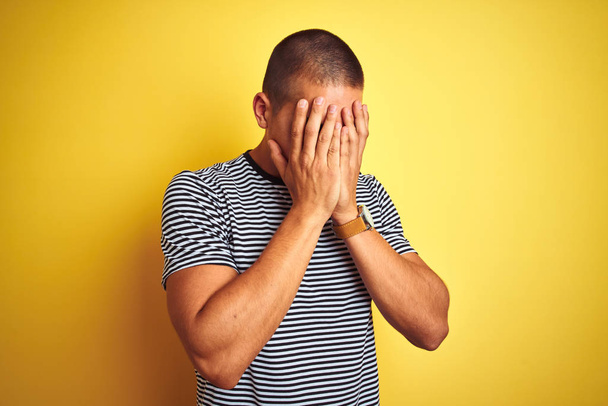 黄色の孤立した背景の上にストライプのTシャツを着た若いハンサムな男は、泣きながら手で顔を覆う悲しい表情をしています。うつ病の概念. - 写真・画像
