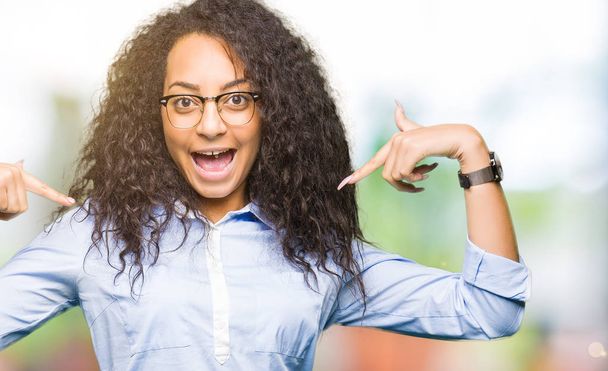 Young beautiful business girl with curly hair wearing glasses looking confident with smile on face, pointing oneself with fingers proud and happy. - Photo, Image