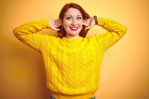 Mulher ruiva bonita usando camisola de inverno em pé sobre fundo amarelo isolado Sorrindo puxando orelhas com os dedos, gesto engraçado. Problema de audição
 - Foto, Imagem