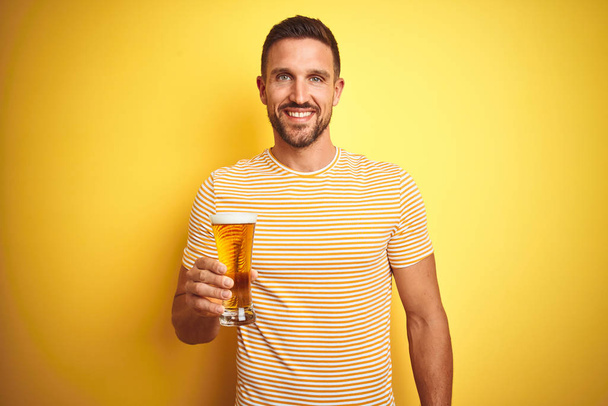 Young handsome man drinking a pint glass of beer over isolated yellow background with a happy face standing and smiling with a confident smile showing teeth - 写真・画像