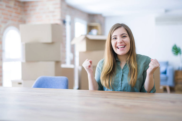 Young woman sitting on the table with cardboard boxes behind her moving to new home celebrating surprised and amazed for success with arms raised and open eyes. Winner concept. - Photo, Image