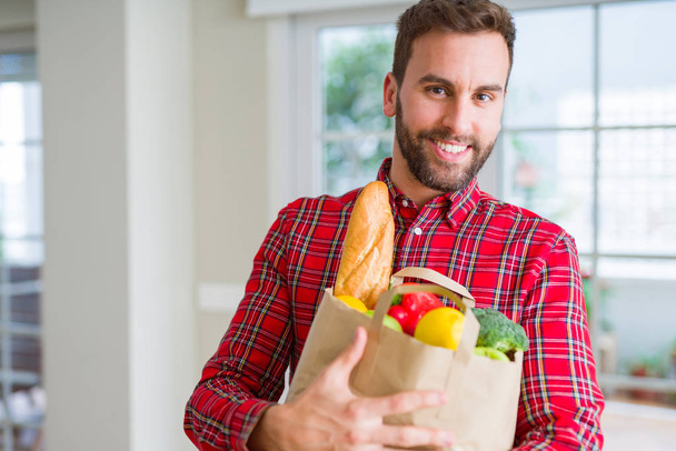 Hombre guapo sosteniendo bolsa de papel llena de comestibles frescos en casa
 - Foto, Imagen