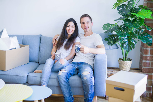 Jeune beau couple assis sur le canapé à boire du café à la nouvelle maison autour des boîtes en carton
 - Photo, image