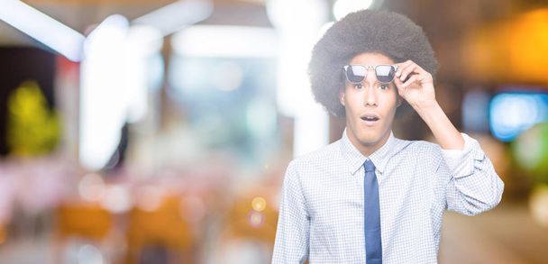 Молодий афроамериканець Ділова людина з афро волосся носити сонцезахисні окуляри боїться і шоковані з сюрпризом вираз, страх і схвильований обличчя. - Фото, зображення