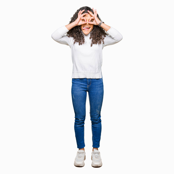 Young beautiful woman with curly hair wearing turtleneck sweater doing ok gesture like binoculars sticking tongue out, eyes looking through fingers. Crazy expression. - Foto, imagen