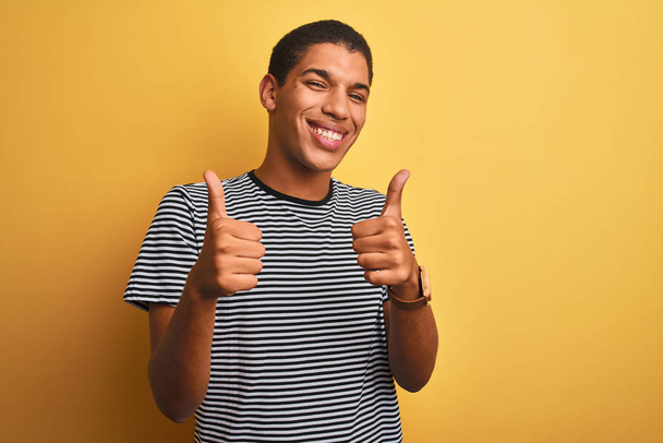Молодой красивый арабский мужчина в футболке с морской полосой, на изолированном желтом фоне знак успеха делает позитивный жест рукой, большой палец вверх улыбается и счастлив. Веселое выражение лица и жест победителя
. - Фото, изображение