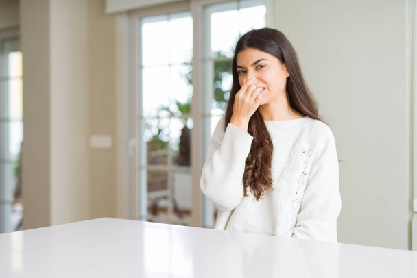 Young beautiful woman at home on white table smelling something stinky and disgusting, intolerable smell, holding breath with fingers on nose. Bad smells concept. - Photo, Image
