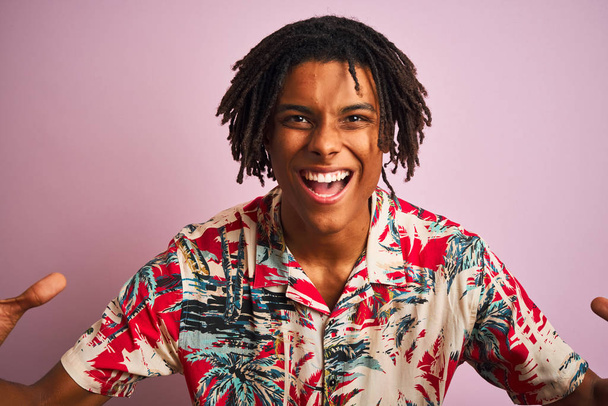 Afro homem com dreadlocks em férias vestindo camisa floral sobre fundo rosa isolado comemorando louco e espantado pelo sucesso com os braços levantados e olhos abertos gritando animado. Conceito de vencedor
 - Foto, Imagem