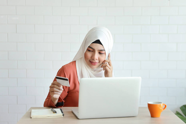 joven musulmana mujer de negocios de compras y pago en línea mediante el uso de cuaderno, teléfono celular y tarjeta de crédito con taza de café en escritorio de madera
 - Foto, imagen