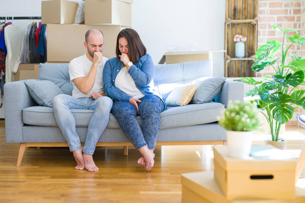 Ein junges Paar, das auf dem Sofa in Pappkartons sitzt und in ein neues Haus zieht, fühlt sich unwohl und hustet als Symptom für Erkältung oder Bronchitis. Gesundheitskonzept. - Foto, Bild