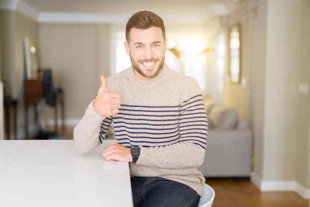 Young handsome man wearing a sweater at home doing happy thumbs up gesture with hand. Approving expression looking at the camera with showing success. - Φωτογραφία, εικόνα