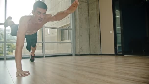 An athletic man with naked torso training in the studio - doing push ups and standing on the one hand and pulling another hand forward - stamina training - Séquence, vidéo