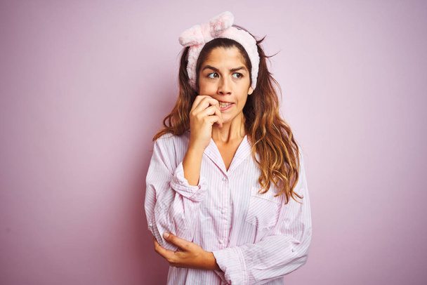 ピンクの孤立した背景の上に立つパジャマを着た若い美しい女性は、口を噛む爪に手を当てて、ストレスと緊張を見ています。不安の問題. - 写真・画像