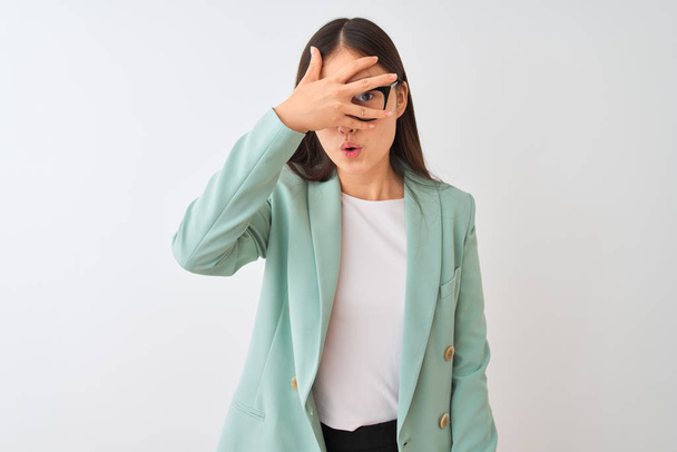 Chinese businesswoman wearing elegant jacket and glasses over isolated white background peeking in shock covering face and eyes with hand, looking through fingers with embarrassed expression. - 写真・画像