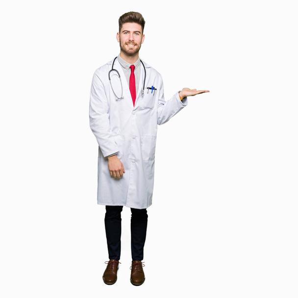 Fiatal jóképű orvos ember visel orvosi kabát, mosolygó, vidám bemutatása és rámutatva a tenyér-ból odaad néz a kamerába. - Fotó, kép