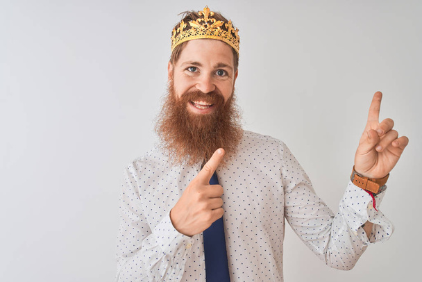Nuori punapää irlantilainen liikemies yllään kruunu kuningas yli eristetty valkoinen tausta hymyillen ja katsoen kameraa osoittaa kahdella kädellä ja sormella sivulle
. - Valokuva, kuva