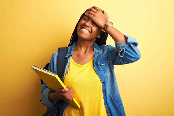 Estudiante afroamericana vistiendo mochila y libro sobre fondo amarillo aislado estresada con la mano en la cabeza, conmocionada con vergüenza y cara sorpresa, enojada y frustrada. Miedo y malestar por error
. - Foto, Imagen