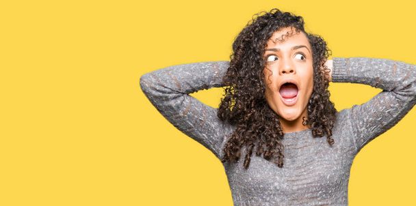 Young beautiful woman with curly hair wearing grey sweater Crazy and scared with hands on head, afraid and surprised of shock with open mouth - Foto, immagini