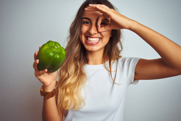 Молодая красивая женщина держит зеленый перец на белом изолированном фоне со счастливым лицом улыбается делает хорошо знак с рукой на глазу глядя сквозь пальцы
 - Фото, изображение