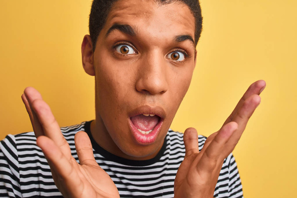 Young handsome arab man wearing navy striped t-shirt over isolated yellow background very happy and excited, winner expression celebrating victory screaming with big smile and raised hands - Photo, Image