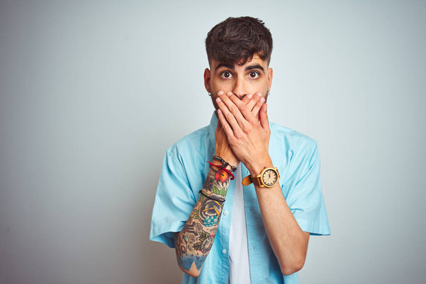 Fiatal férfi tetoválás visel kék inget állt át elszigetelt fehér háttér döbbenve, amely szája kézzel a hiba. Titkos koncepció. - Fotó, kép