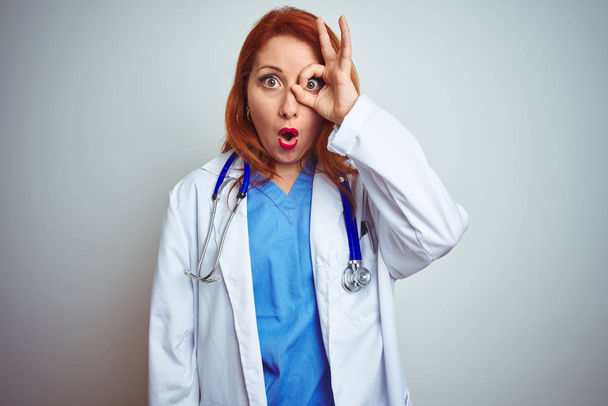 Молодая рыжая женщина-врач, использующая стетоскоп на белом изолированном фоне, делает нормальный жест, шокированный удивленным лицом, глазами, смотрящими сквозь пальцы. Неверное выражение
. - Фото, изображение